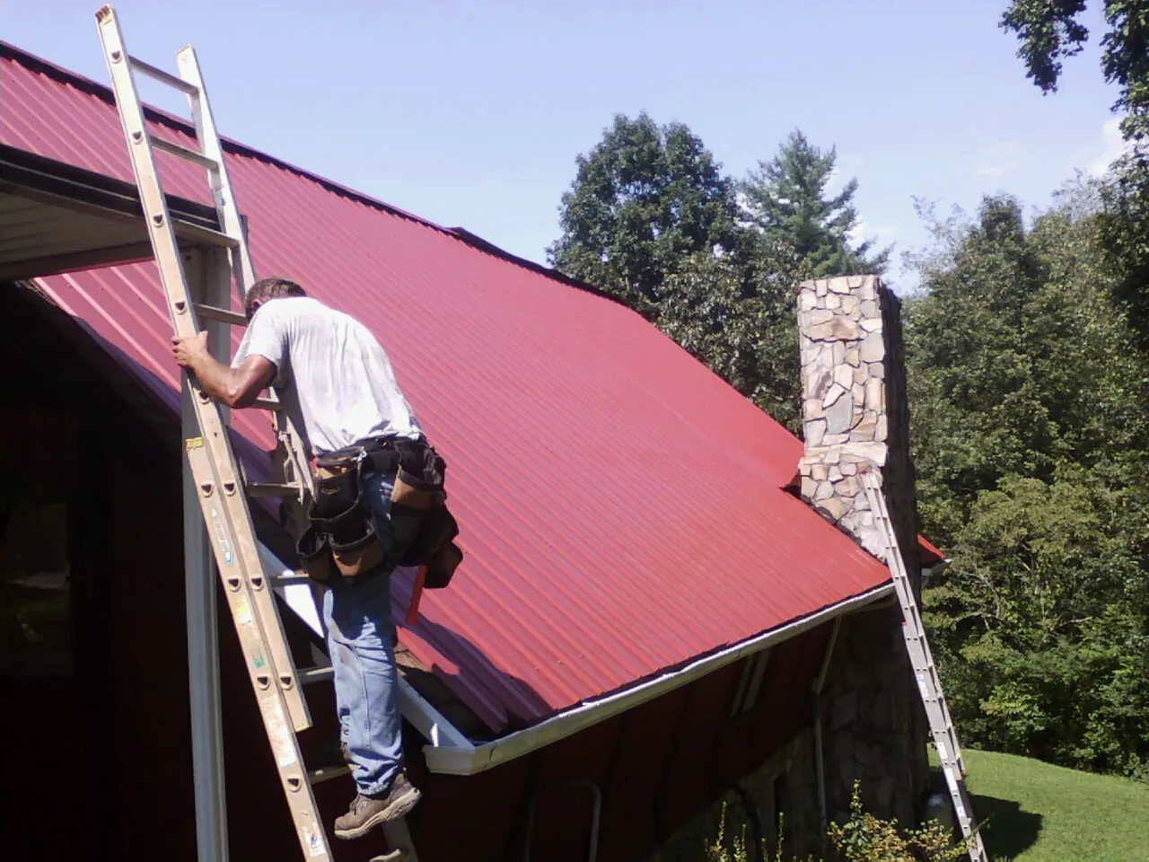 Roofer on a ladder beside a red metal roof and stone chimney — Soffit & Fascia Repair by Roofing Brothers in Rayville